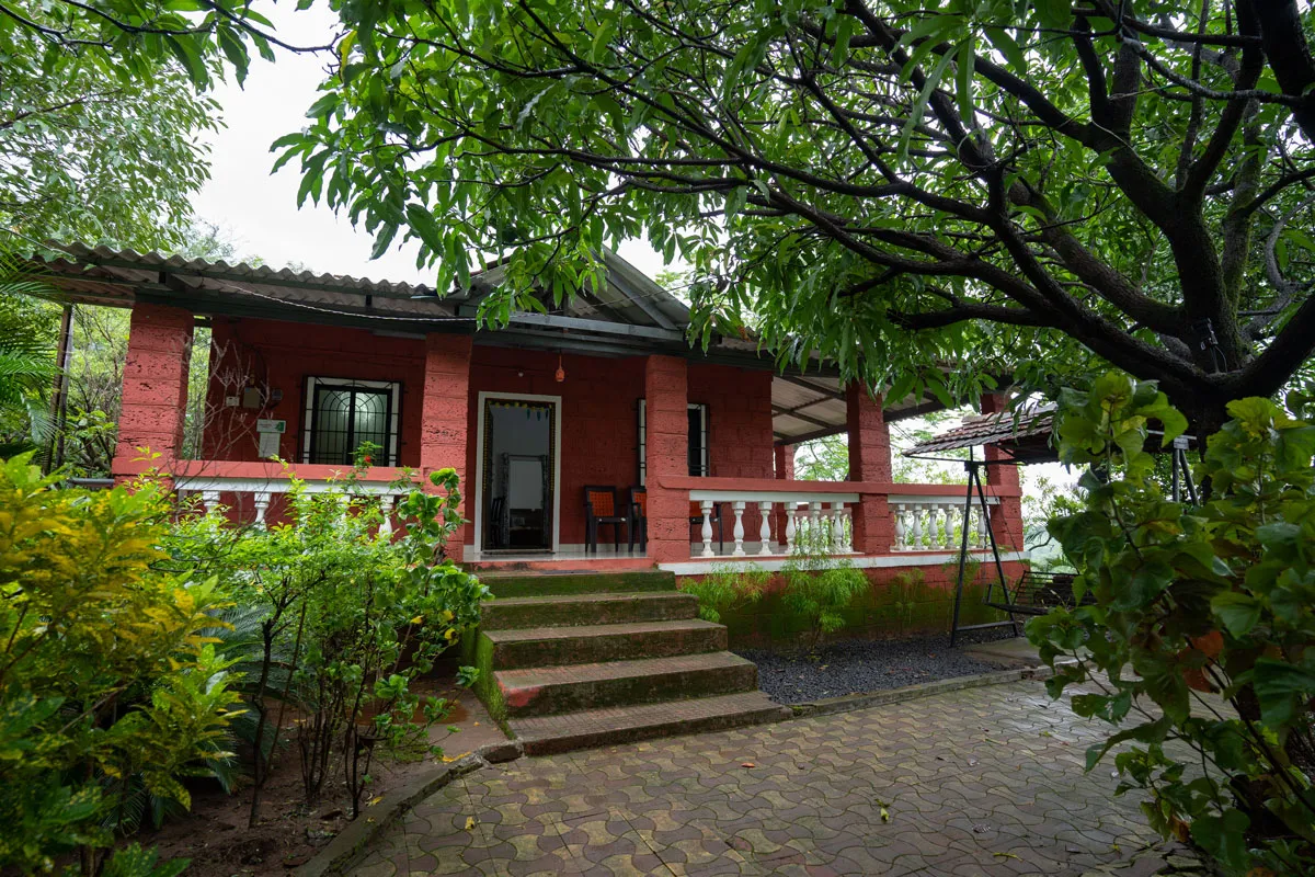 Red building with a covered veranda and white railing, surrounded by lush green trees and plants.
